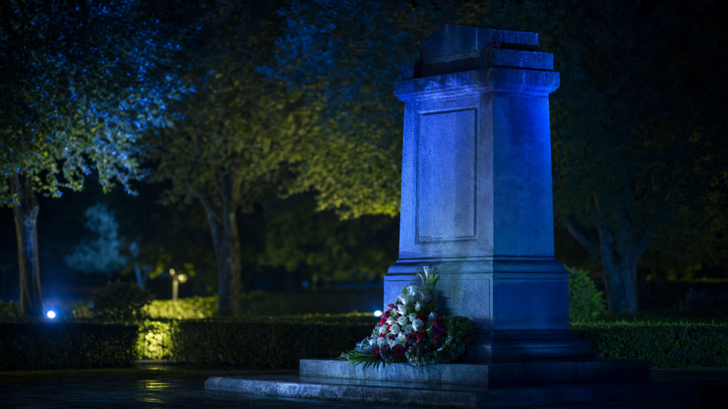 Stone monument illuminated in blue light at night with flowers at the base